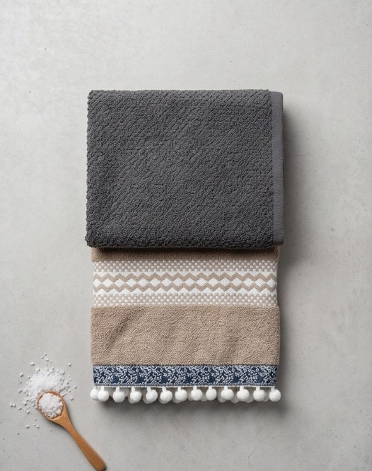 Two folded towels, one gray and one with a pattern and pom-poms, on a light gray surface.