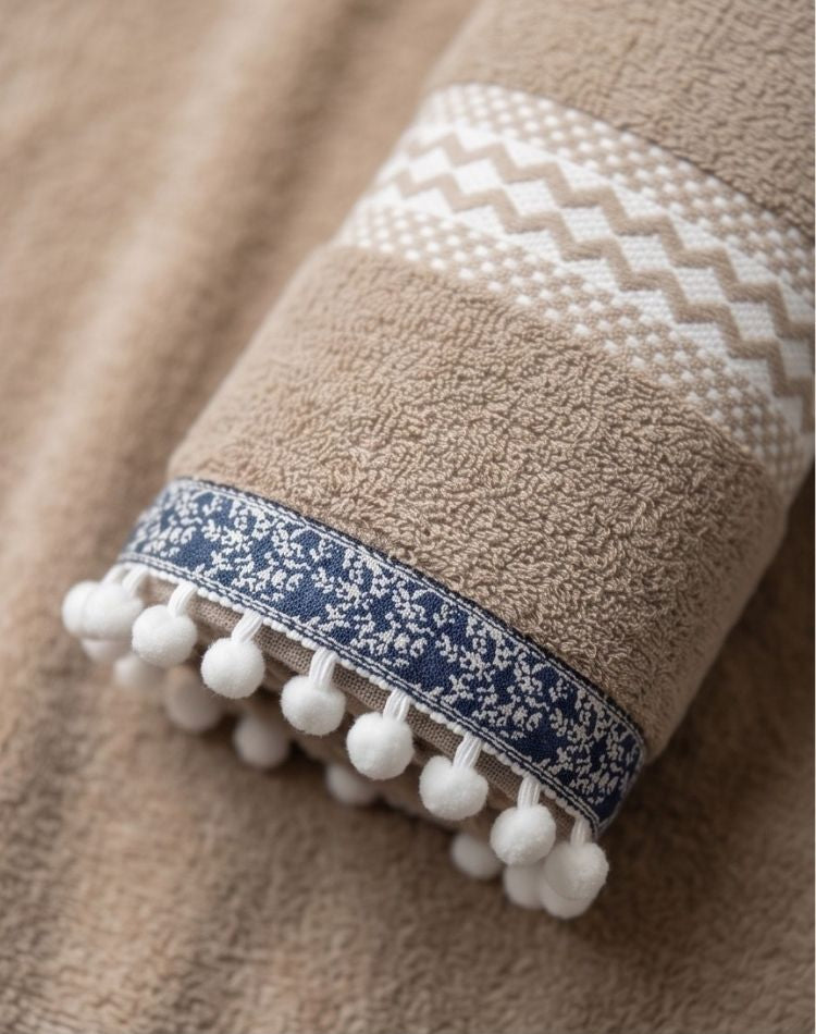 Brown towel with white geometric pattern and blue border with white pom-poms on a matching brown background.
