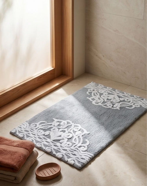 Gray bath mat with white lace pattern on a wooden floor next to a window.