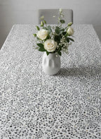 A white tablecloth with a dense black floral and leaf pattern, styled with a white ceramic vase holding white roses and green foliage in the center, with a gray chair visible in the background.